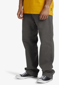 Pantaloni gri din bumbac, cu o croială lejeră, alături de o cămașă galbenă. Pantofi sport negri cu guler înalt și tălpi albe completează ținuta.