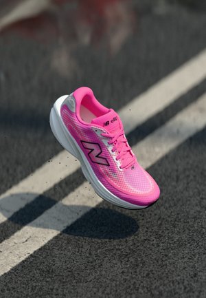 Felle roze New Balance hardloopschoen zwevend boven een asfaltweg met witte rijstrookmarkeringen, vuildeeltjes zichtbaar onder de zool.