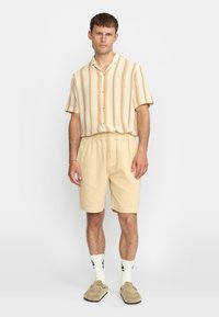 Lichtgekleurde korte mouwen overhemd met verticale strepen; beige elastische shorts; witte sokken met zwarte accenten; bruine instappers.