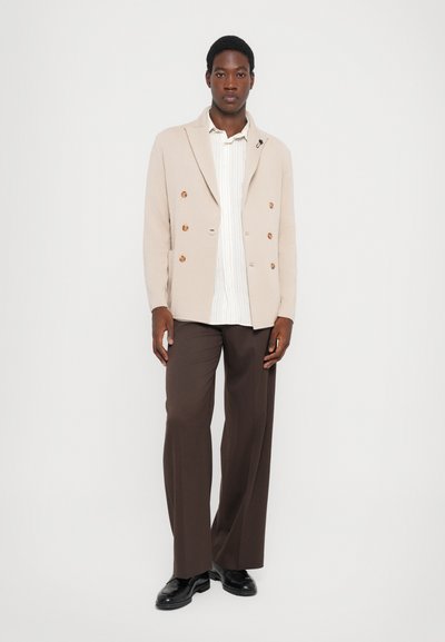 Beige Doppelreiher-Blazer mit Holzknöpfen über einem weißen gestreiften Hemd, kombiniert mit dunkelbraunen weiten Hosen und schwarzen Schuhen.