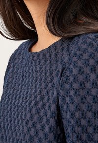 Top blu navy con tessuto testurizzato e motivi floreali ricamati, scollatura rotonda e maniche corte. Vista ravvicinata dei dettagli del design.