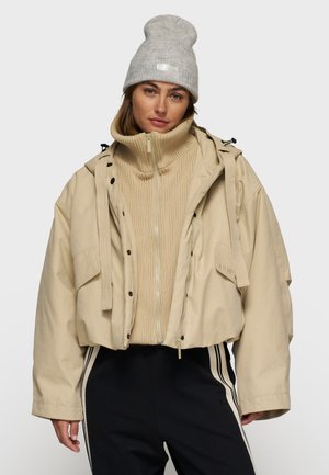 Beige cropped jack met ritssluiting aan de voorkant, grote zakken, verstelbare capuchon en zwarte accenten boven een tan geribbelde trui en donkere broek. Grijze beanie.