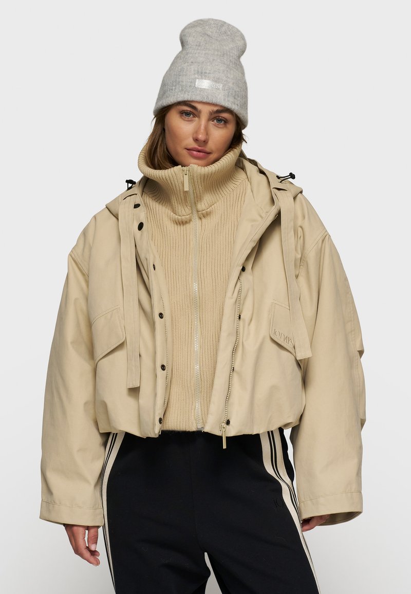 Beige cropped jakke med frontlynlås, store lommer, justerbar hætte og sorte detaljer over en tan ribbestrikket trøje og mørke bukser. Grå hue.