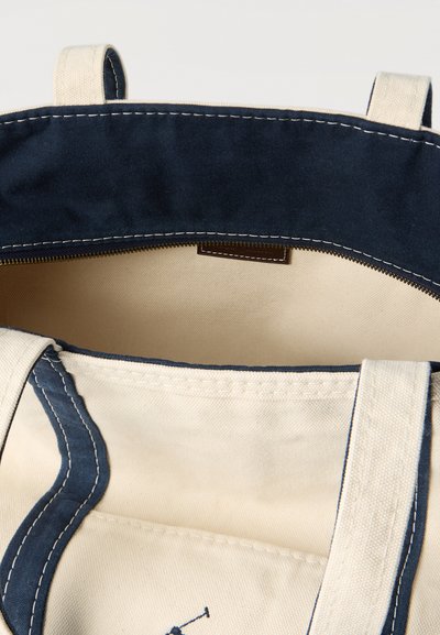 Τσάντα tote από καμβά με περιθώριο σε σκούρο μπλε, μπεζ εσωτερικό και διπλές λαβές, εμφανιζόμενη ανοιχτή με ορατό φερμουάρ στο επάνω άκρο.