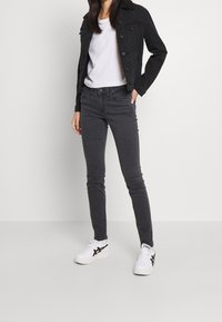 Chaqueta de mezclilla negra sobre una camiseta blanca, combinada con jeans ajustados grises y zapatillas blancas con detalles negros. Estilo simple y ceñido.