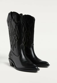 Čierne kožené kovbojské topánky s špicatými špičkami, zložitým prešívaním a nízkym blokovým podpätkom. Majú vysoký nátepník a dekoratívne akcenty.