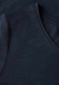 Detailní záběr na tmavě modrou látku se měkkou texturou, která má výstřih do V a jemné prošití podél okrajů.