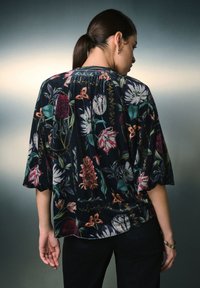 Blusa floreale in tessuto scuro con grandi fiori colorati e motivi fogliari. Maniche a sbuffo e dettaglio con scollo arricciato.