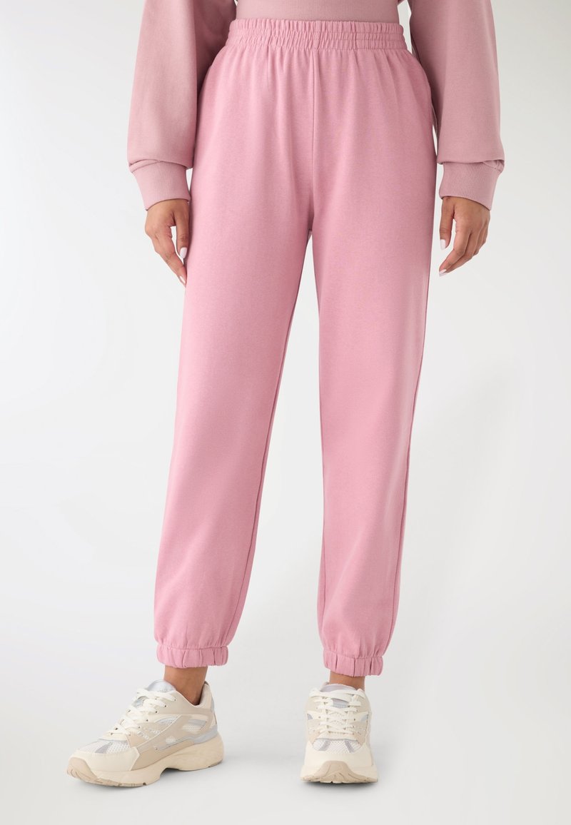 Persona che indossa pantaloni rosa a vita alta con polsini elasticizzati e sneakers bianche chunky, in piedi su uno sfondo chiaro e uniforme.