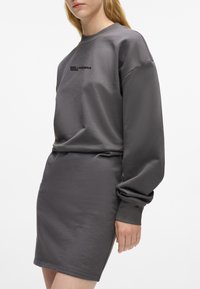 Kvinne iført en grå langermet sweatshirtkjole med "KARL LAGERFELD JEANS"-logo på brystet, stående mot en hvit bakgrunn.