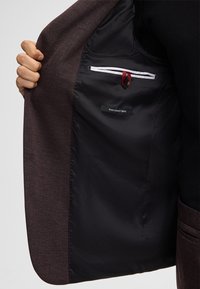 Giacca sportiva marrone scuro con un interno nero liscio, dotata di una tasca con zip e un'etichetta che recita "miglior tessuto elasticizzato". Accenti rossi visibili.