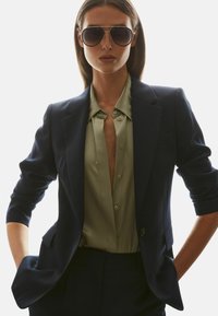 Kvinde iført store mørke solbriller, navy blazer med opkørte ærmer og delvist åbnet olivengrøn skjorte, hænderne i lommerne.