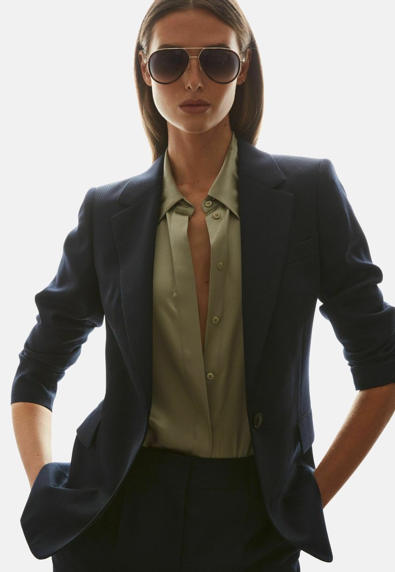 Kvinde iført store mørke solbriller, navy blazer med opkørte ærmer og delvist åbnet olivengrøn skjorte, hænderne i lommerne.