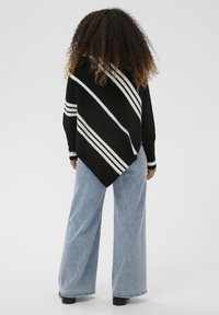 Asmuo su garbanotais plaukais, vilkintis juodai baltą raižytą asimetrinį pončą, šviesiai mėlynas platėjantys džinsus ir juodus batus, stovi atsisukęs atgal.