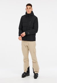 Schwarze gepolsterte Jacke mit Kapuze, mit Reißverschlusstaschen und elastischen Bündchen; kombiniert mit beigen Skihosen und schwarzen Skischuhen.