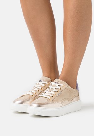 Béžové a kovově zlaté tenisky s bílými tkaničkami nošené na bosých nohou na jednobarevném bílém pozadí.