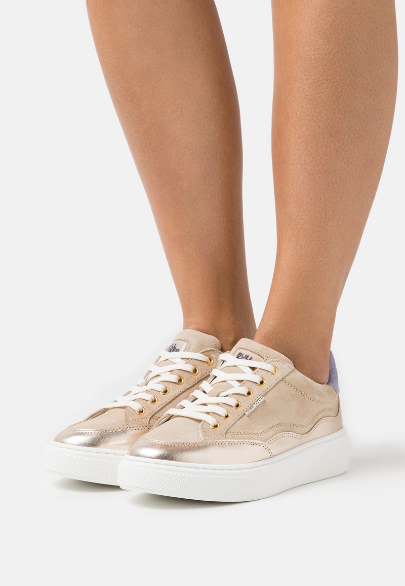 Baskets beige et or métallique avec des lacets blancs portées pieds nus sur un fond blanc uni.