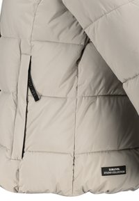 Helle graue gesteppte Pufferjacke mit Seitentasche, ausgestattet mit einem schwarzen Ziehband und einem schwarzen Etikett mit der Aufschrift "Sublevel Studio Collection".