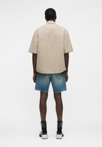 Hombre con camisa de manga corta a cuadros beige, pantalones cortos de mezclilla con dobladillo deshilachado y botas negras de tobillo con tacones gruesos, de espaldas sobre fondo liso.
