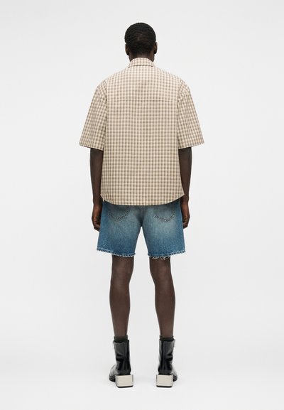 Homme portant une chemise à manches courtes beige à carreaux, un short en jean à ourlet effiloché et des bottines noires à talons épais, de dos sur un fond uni.