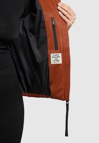 Ruda striukė su užtrauktuku, lygiu išoriniu paviršiumi ir juodu vidiniu pamušalu. Yra etikėtė su užrašu ir audinio detalės.