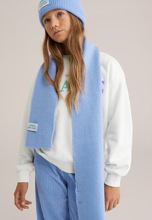 Világoskék kötött sapka és hozzáillő, bordázott mintájú sál, fehér pulóver és világoskék csíkos nadrág felett viselve. A sálon címke található.