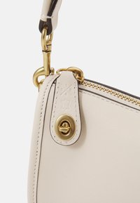 Sac à main en cuir beige clair avec quincaillerie dorée, doté d'une fermeture éclair et d'une étiquette en cuir décorative embossée avec un logo et des détails de design.