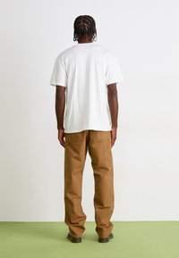 Man with braided hair stands facing away, wearing a white t-shirt, brown cargo pants, and black shoes on green floor.