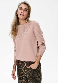 Kobieta w jasnoróżowym swetrze dzianym z teksturowymi wzorami oraz w spodniach w panterkę z brązowym paskiem, stojąca z jedną ręką w kieszeni.
