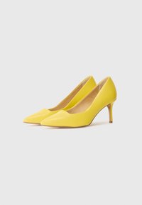Paire d’escarpins jaunes à bout pointu avec talons aiguilles de hauteur moyenne sur fond blanc uni.