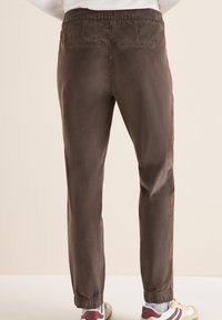 Bruine elastische broek met achterzakken en taps toelopende enkels. Glad materiaal met een subtiele glans. Gestyled met sneakers.
