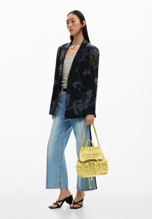 Mujer con blazer oscuro estampado de flores, top gris, vaqueros de pierna ancha, sandalias negras de tacón, collar plateado, sosteniendo un bolso amarillo con lazos.