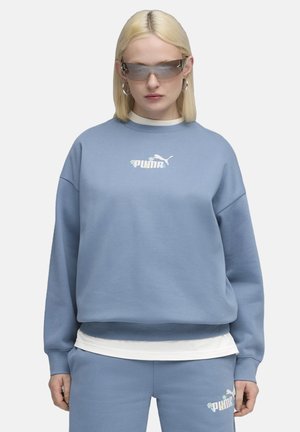 Sweatshirt bleu clair à coupe décontractée et col rond, avec un logo Puma blanc. Superposé avec une chemise intérieure blanche, assorti avec un pantalon. 