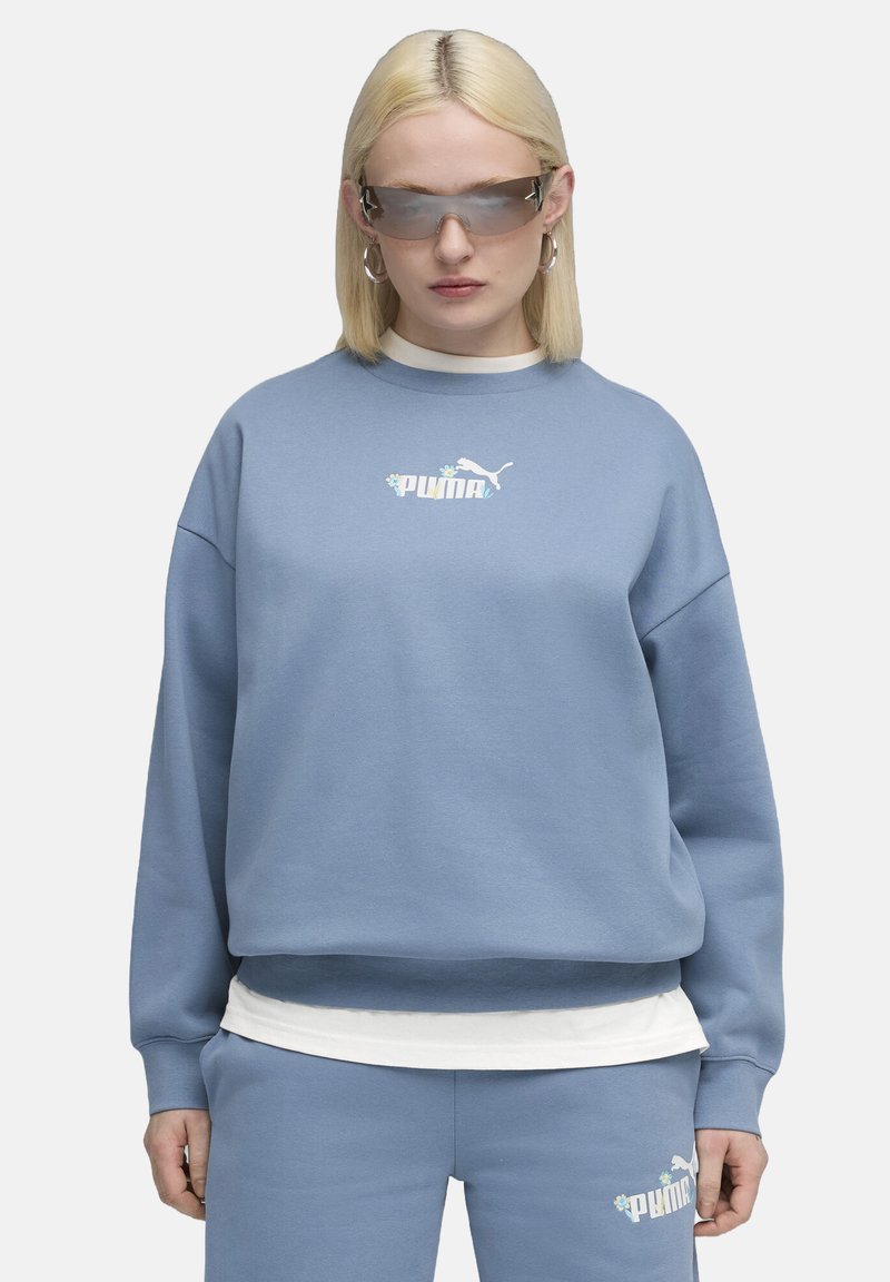 Svetlomodrá mikina s voľným strihom a okrúhlym výstrihom, s bielym logom Puma. Preložená s bielou vnútornou košeľou, skombinovaná s ladiacimi nohavicami.