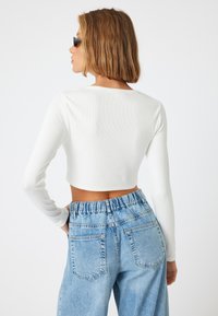 Weißes, geripptes Langarm-Crop-Top kombiniert mit hellblauen, hochgeschnittenen Jeans aus Denim mit elastischem Bund und Gesäßtaschen.