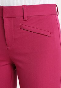 Pantalon magenta à texture lisse, avec une coupe ajustée, des passants pour ceinture et une poche passepoilée unique sur le côté droit.