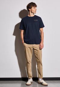 Mörkblå bomull t-shirt med kort ärm och tryckt logotyp, kombinerad med beige chinos och vita sneakers med bruna detaljer.