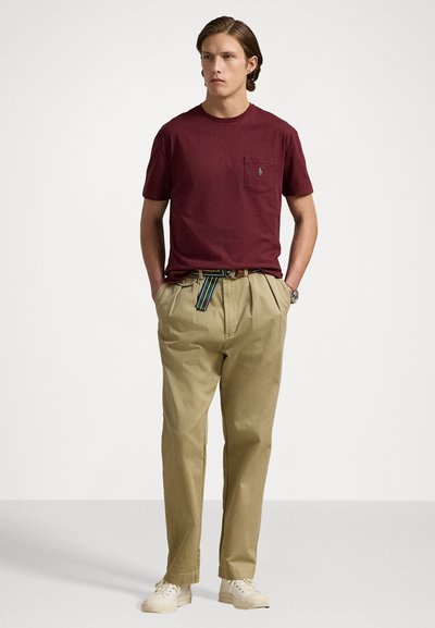 Vinröd bomullströja med framficka och logotyp, kombinerad med beige byxor och ett flerfärgat randigt bälte. Vita sneakers fulländar outfiten.
