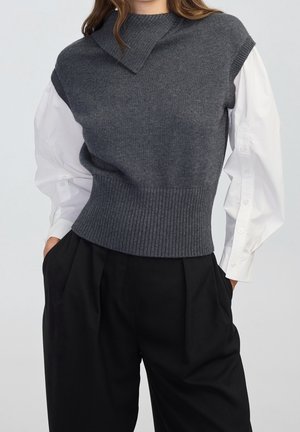 Vrouw draagt een donkergrijs mouwloos gebreid vest over een wit blouse met lange mouwen, gecombineerd met losse zwarte broek, staand met handen in de zakken.