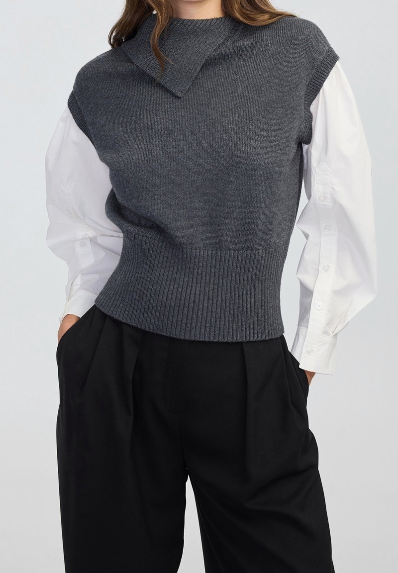 Femme portant un gilet sans manches en maille gris foncé sur un chemisier blanc à manches longues, associée à un pantalon noir ample, debout les mains dans les poches.