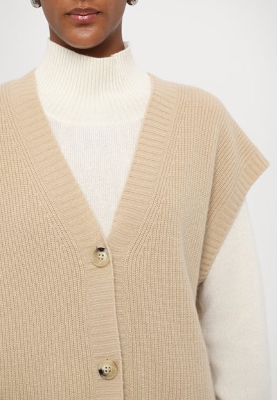 Beige strikket vest med dyb V-udskæring og korte ærmer, lukket med en enkelt knap foran, lagt over en cremefarvet turtleneck sweater.