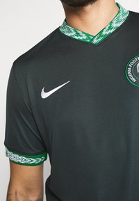 Hombre vestido con una camiseta oscura verde de la Federación de Fútbol de Nigeria, con un logo blanco de Nike y un cuello y puños de manga azul con patrones verdes.