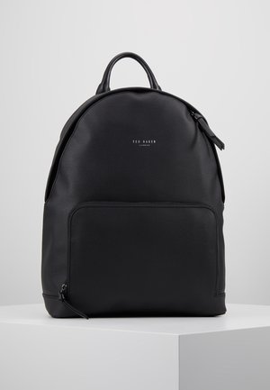 Czarny skórzany plecak Ted Baker z górnym uchwytem, przednią kieszenią na zamek oraz wytłoczonym logo marki na gładkiej powierzchni, stojący na białym pedestale.