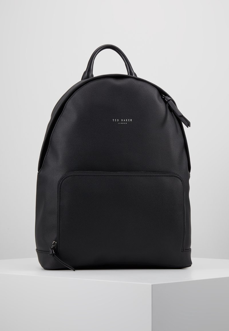 Sac à dos en cuir noir Ted Baker avec poignée supérieure, poche avant zippée et logo de marque embossé sur une surface lisse, posé sur un socle blanc.