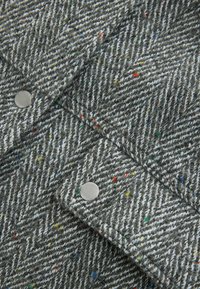 Harmaa herringbone-kangas, jossa on monivärisiä pilkkuja. Varustettu metallipainonapeilla. Tekstuurin kerrotaan olevan pehmeä ja teksturoitu, jossa on erottuvia valkoisia kuituja.