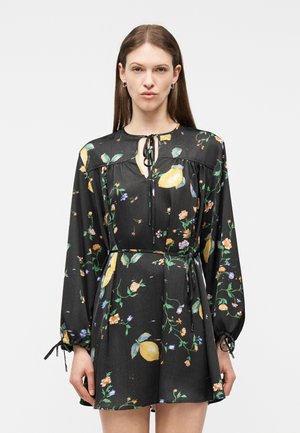 Nő fekete, hosszú ujjú mini ruhában, amely sárga citrom- és virágmintás, nyakán megkötős és derékban övvel, egyszínű háttér előtt áll.