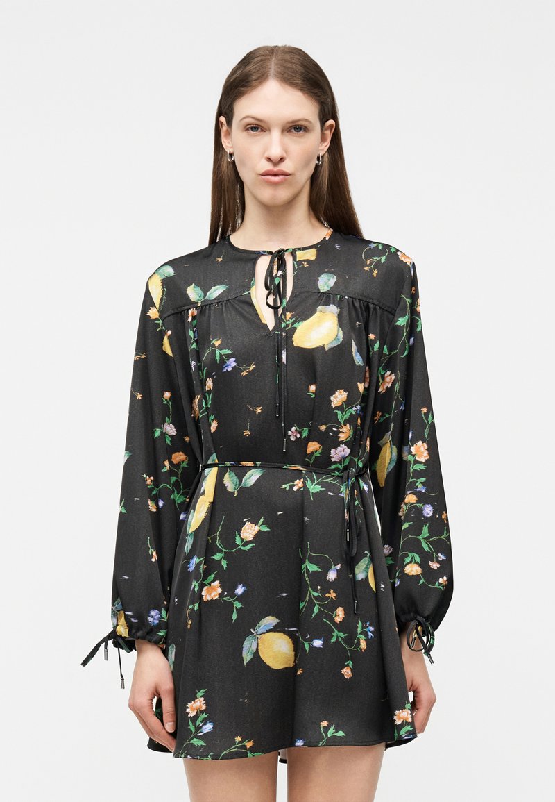 Γυναίκα που φοράει μαύρο μίνι φόρεμα με μακριά μανίκια, κίτρινο λεμονόχρωμο και λουλουδένιο μοτίβο, κορδόνι στο λαιμό και ζώνη στη μέση, στέκεται μπροστά σε ένα απλό φόντο.