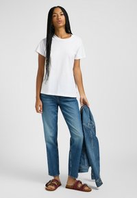 T-shirt bianco in cotone, vestibilità rilassata, maniche corte. Abbinato a jeans blue straight leg e sandali marroni con fibbie decorative. Tiene in mano una giacca di denim.