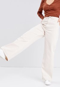 Beige vide ben-jeans lavet af bomuld, med høj talje, frontlommer og en glat tekstur. Sammen med hvide sneakers.