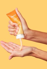 Die Murad Vitamin C Gesichtscreme in einer orangefarbenen Tube wird ausgequetscht, wodurch ein cremiges, gelbliches Produkt in eine Hand released wird. Der Hintergrund ist einfarbig gelb.